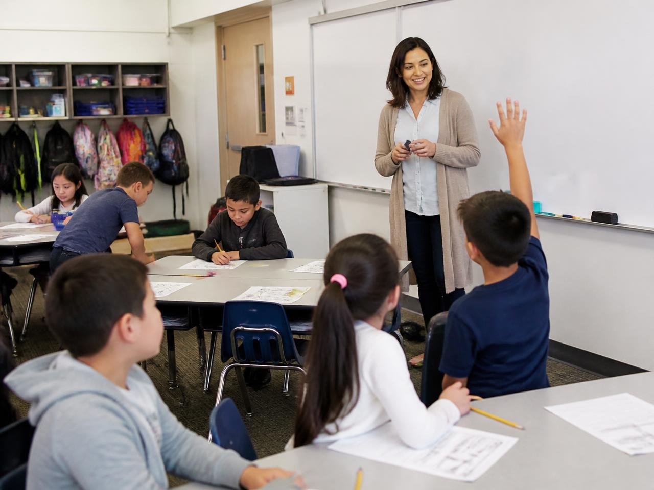 An elementary classroom with a teacher at the board and students raising hands at their desks