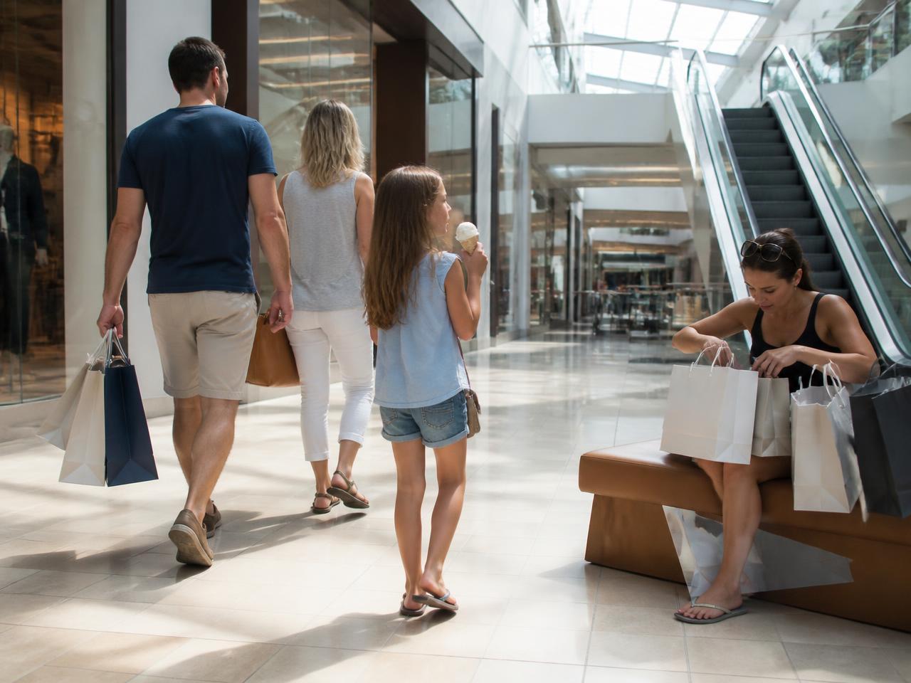 A shopping mall walkway with a family carrying bags, a girl eating ice cream and a woman resting on a bench near the escalator