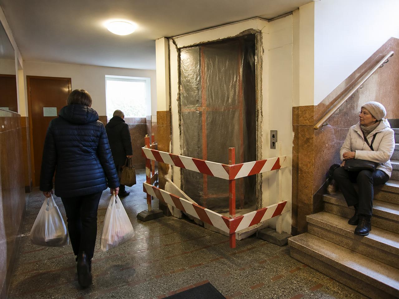 An apartment lobby with the elevator sealed off for replacement while residents carry bags to the stairs and an older woman rests on the steps